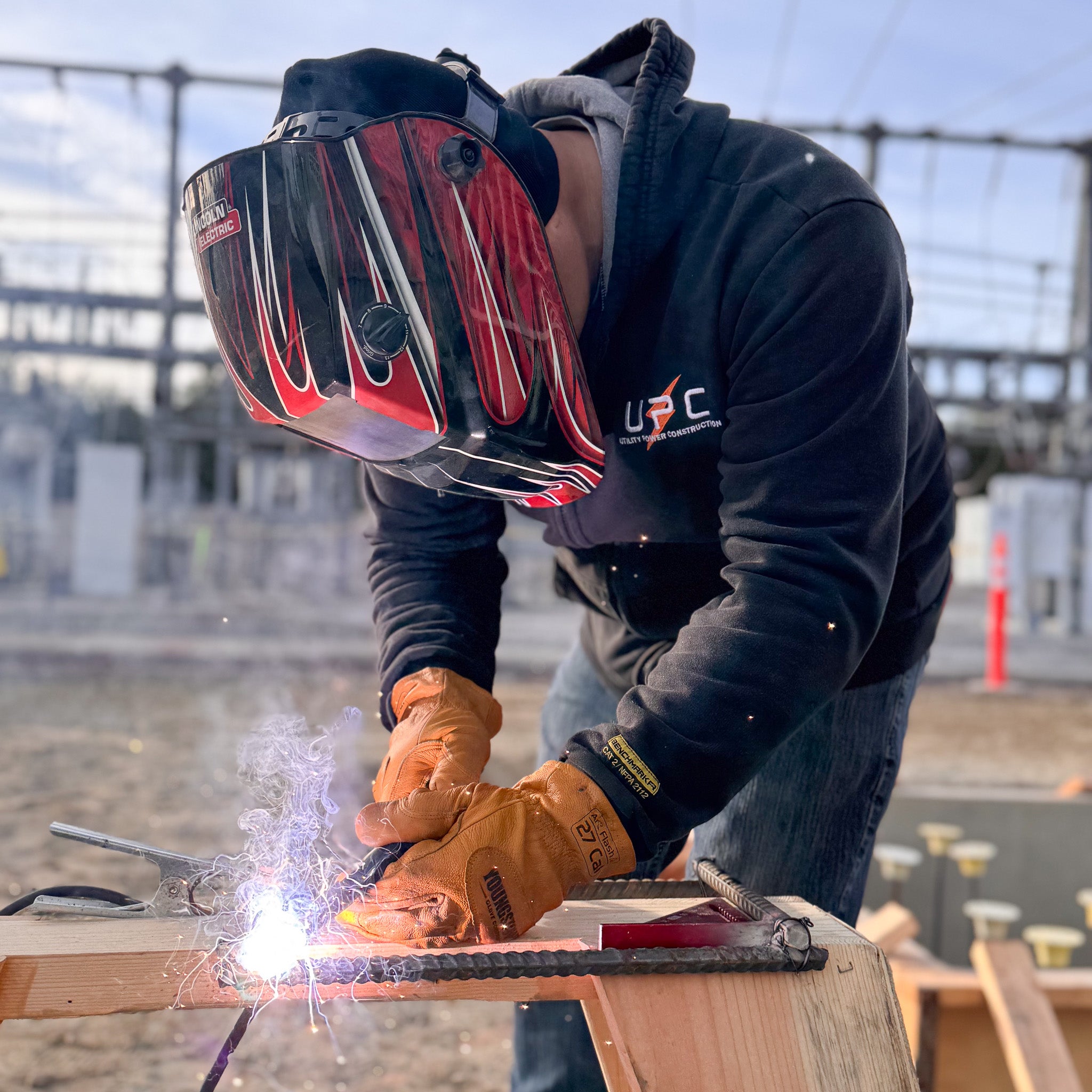 A welder wearing a black hoodie, a red and black flame-designed welding helmet, and protective orange gloves is focused on welding a metal rebar onto a wooden framework. Bright sparks and smoke rise from the point of contact as the worker leans forward, carefully controlling the weld. The background features an industrial construction site with electrical infrastructure and metal frameworks under a clear sky.